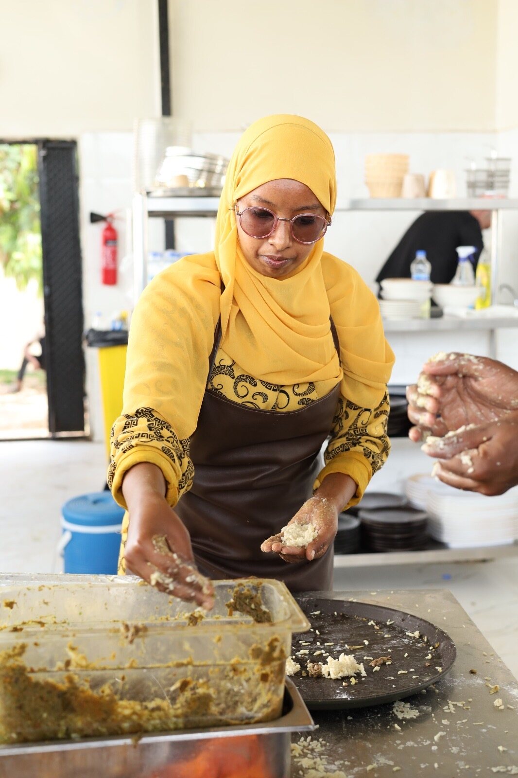 Cook in a yellow hijab and apron shaping food at a kitchen prep station